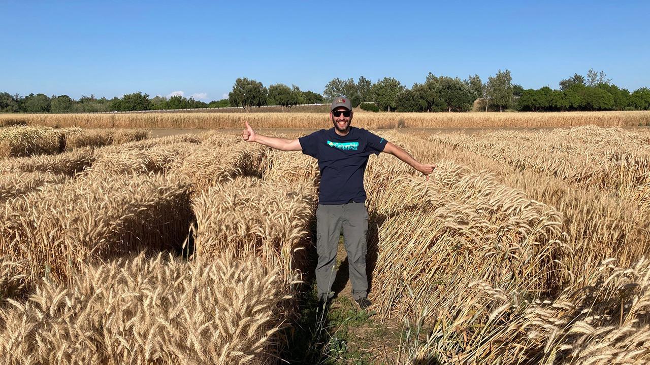 A man smiling and wearing a hat, standing between two fields of light brown grass. Grass in the plot on the right is taller and noticeably bent over.