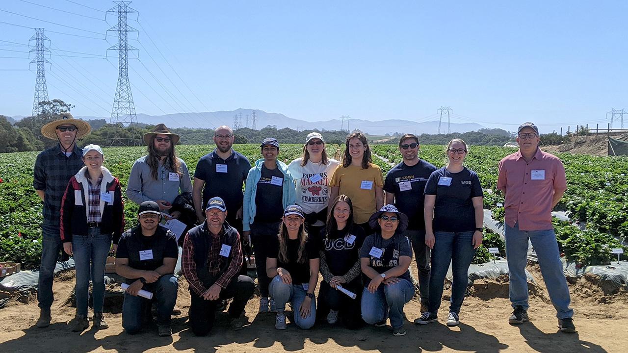 A large group of people standing in front of low furrows, with low mountains and blue sky in the background.