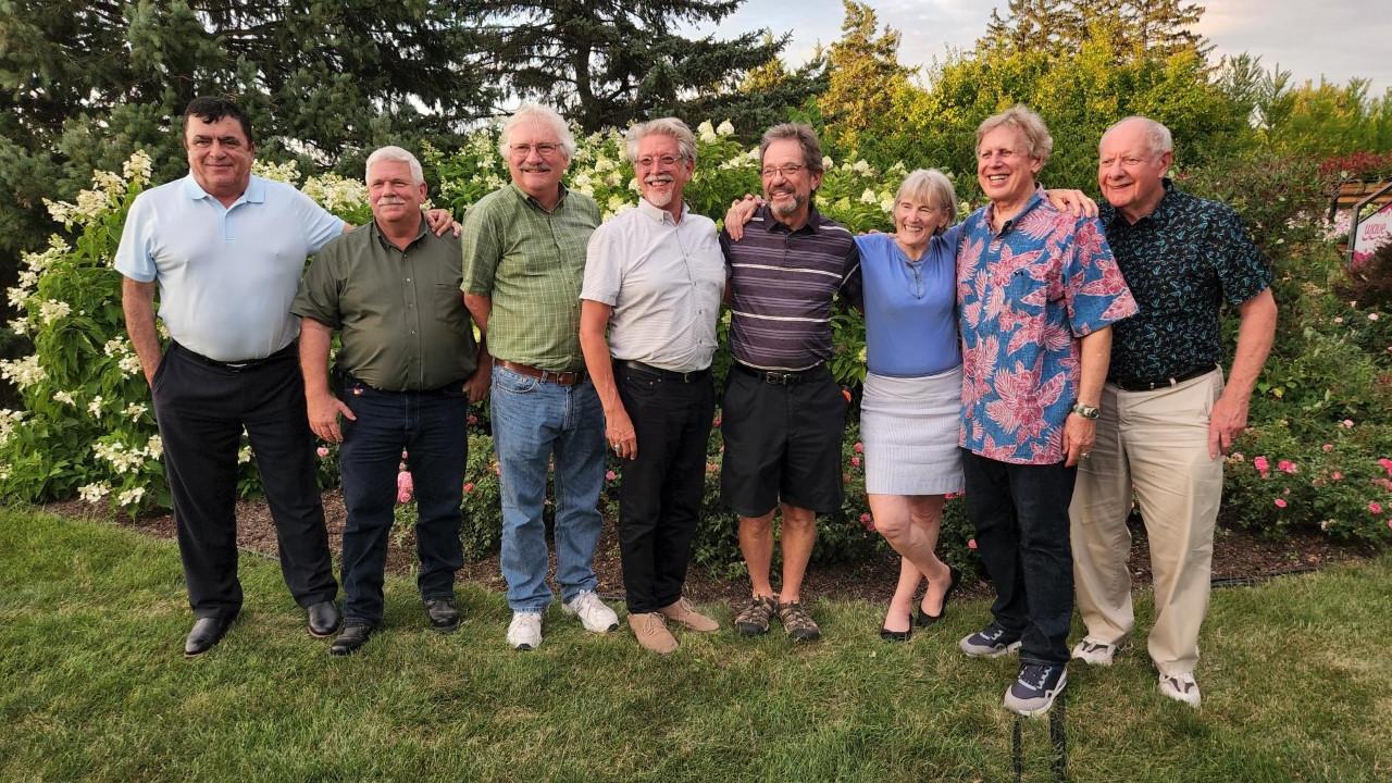 Eight men and one woman stand in a row, smiling, against a backdrop of trees, lawn and cloud-studded sky