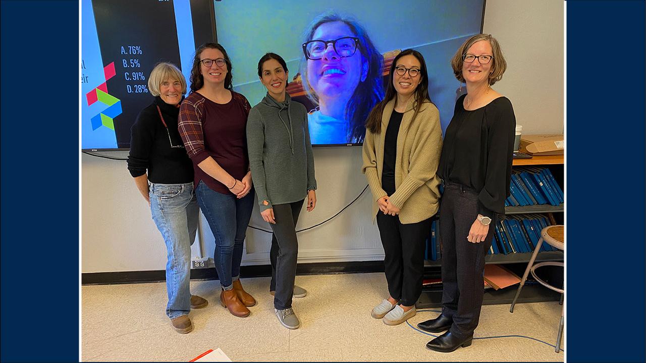 Five women standing in a room. A sixth woman appears on a large TV screen behind them.