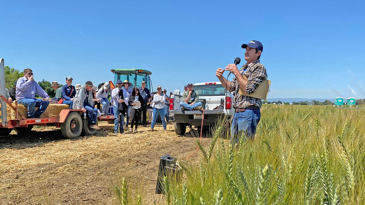 People are gathered at the edge of a field, listening to a man speaking with a microphone