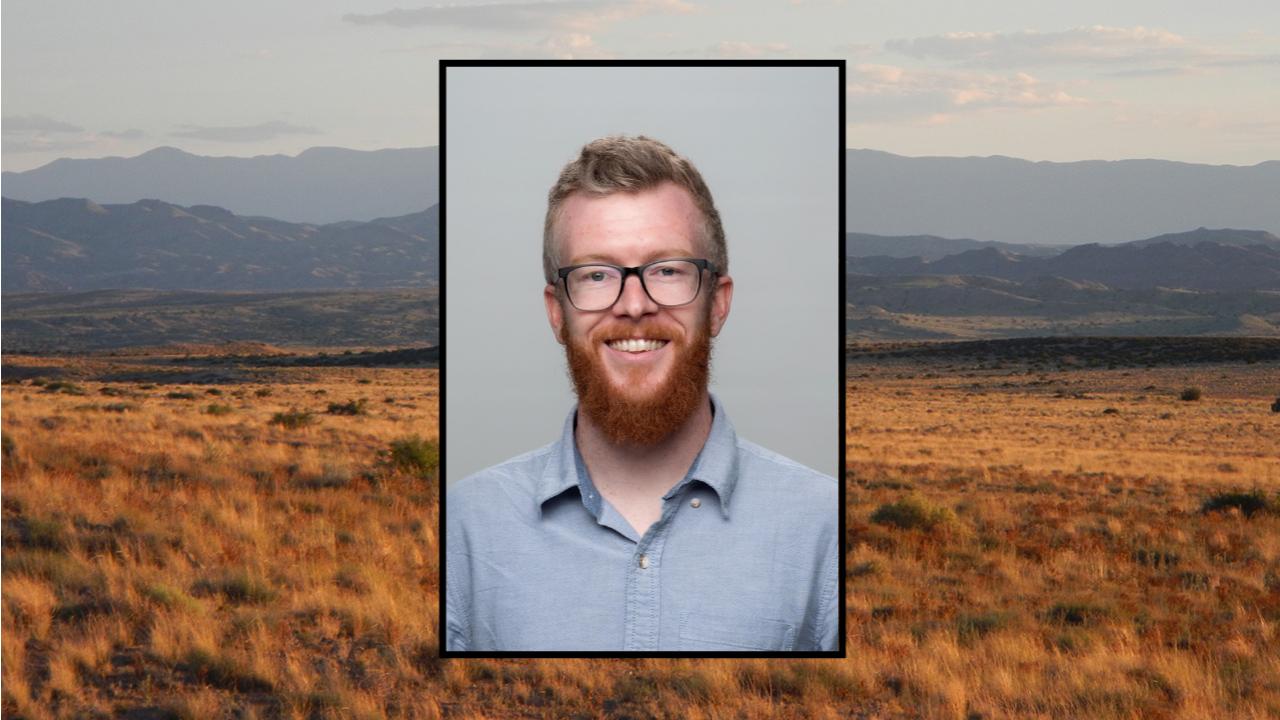 Head-shot of a man, with background photo showing a broad landscape of grown grassland. At the far horizon are hazy blue mountains and gray-blue cloudy sky