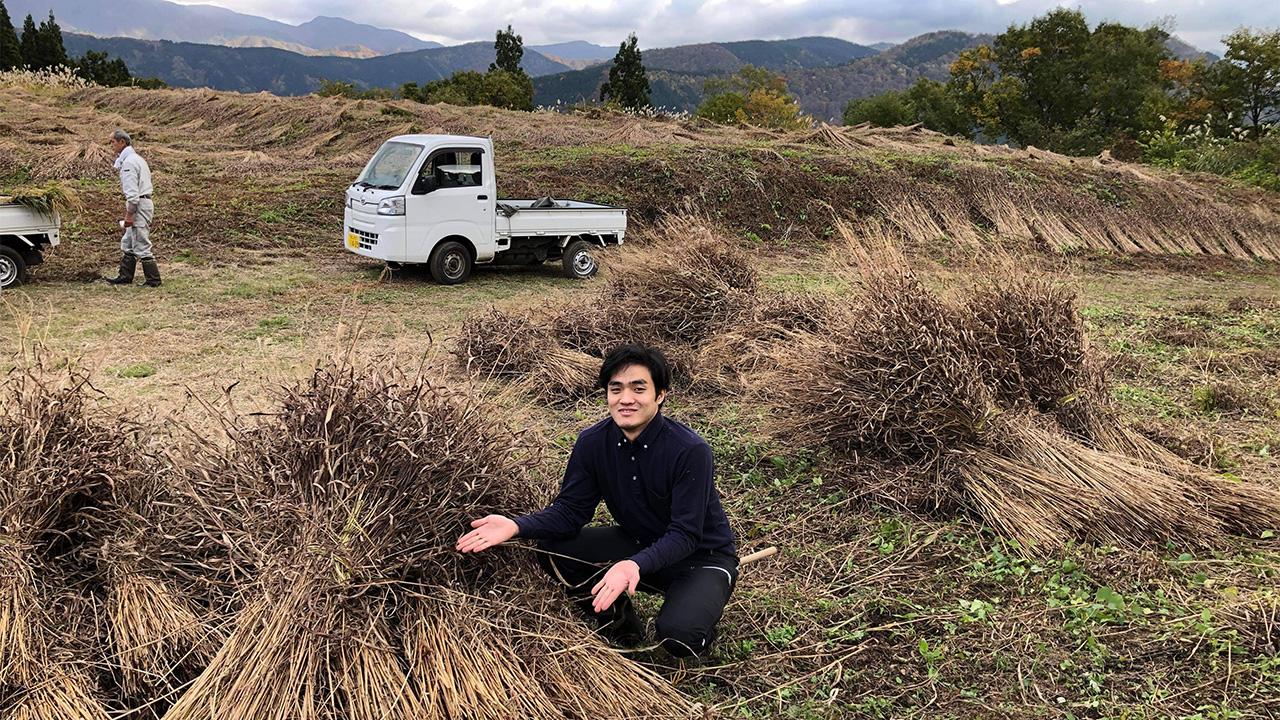 A young man at the edge of a field. Bundles of grass are heaped along the edge, A small truck behind him, and mountains on the near horizon.