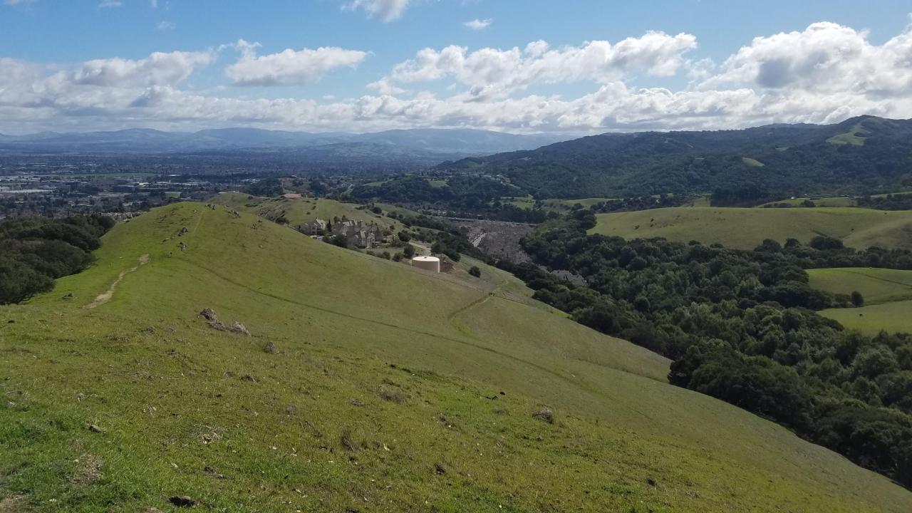 A rolling landscape view from atop a hill looking down into the valley. Trees dot the area with clouds near the horizon.
