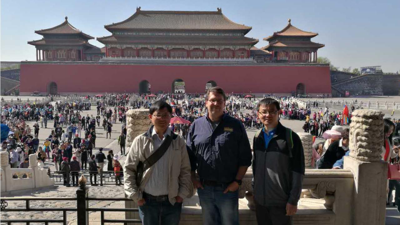 Three men standing in front of a large open square. Buildings in background of Asian architecture.