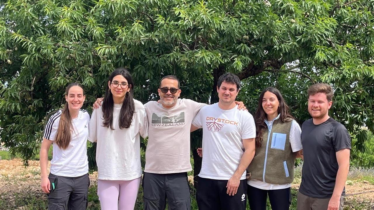 A group of six people standing together outdoors in front of a green tree.