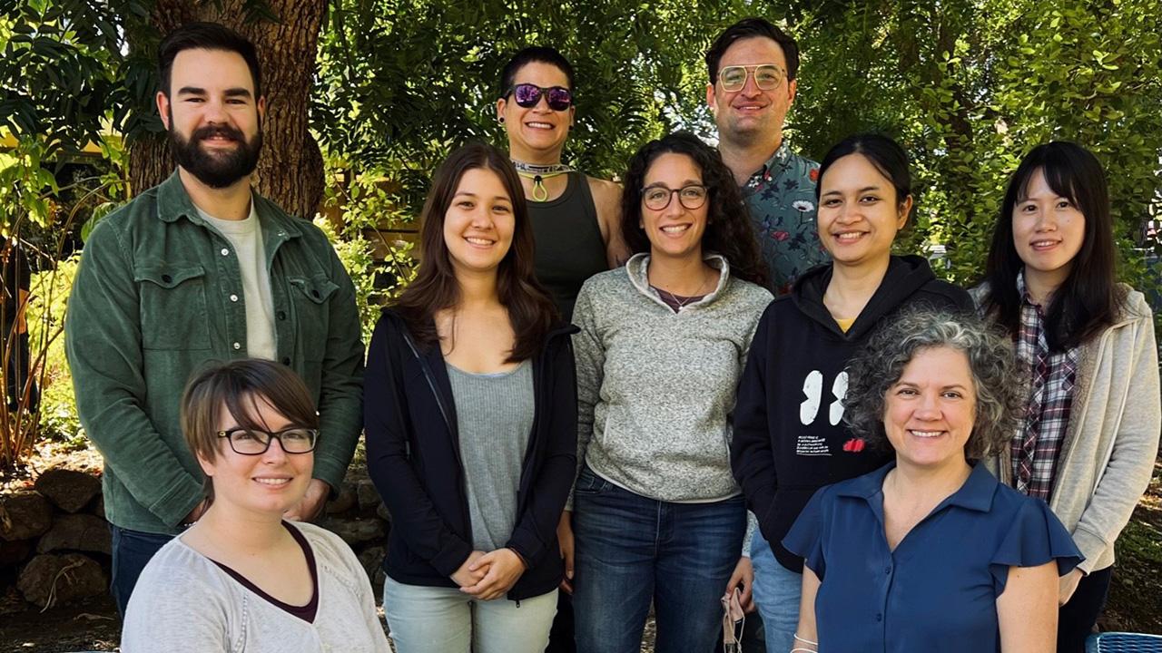 A smiling woman sits in the lower right, while seven younger people stand near her, with lush green trees in the background.