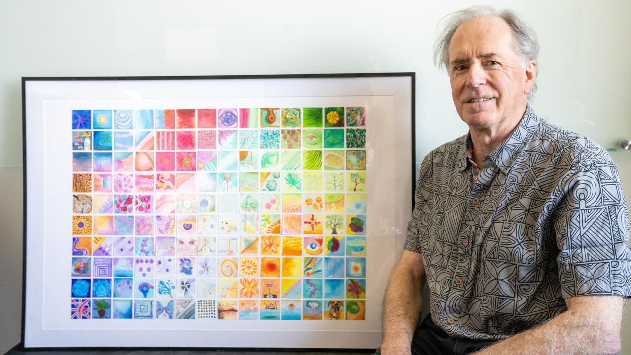 An older man standing next to a large and colorful framed piece of art.
