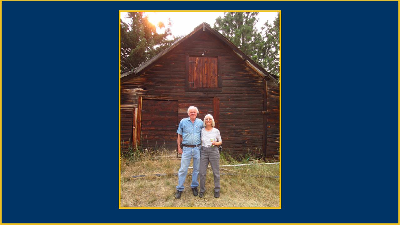 An older man and woman standing in front of an old-looking barn, smiling. The man has one arm around the woman.