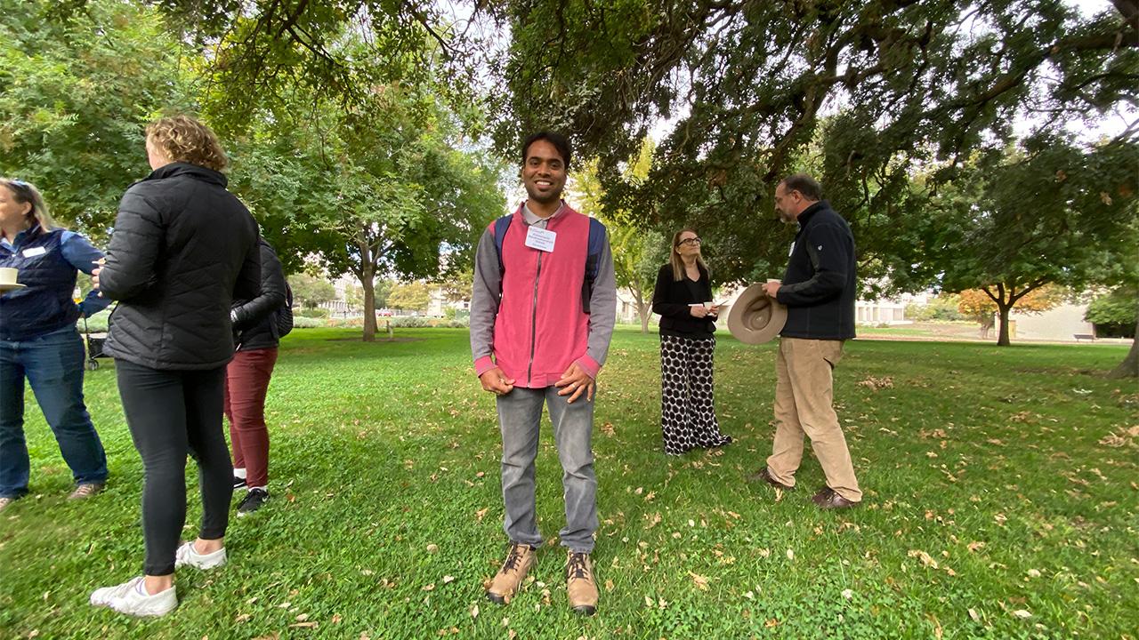 Man standing on grass under large spreading tree, with a few other people nearby chatting.