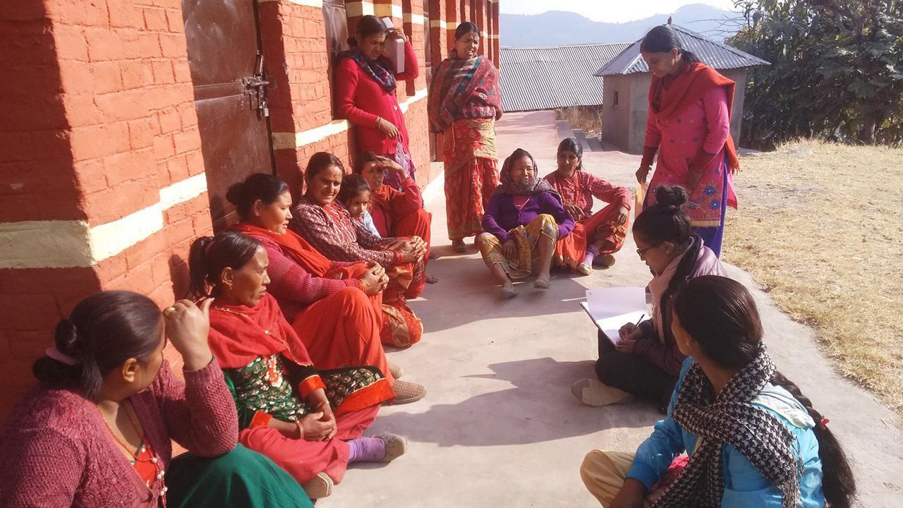 Women wearing colorful red garments, sitting by rustic buildings with the view of high mountains in the background.