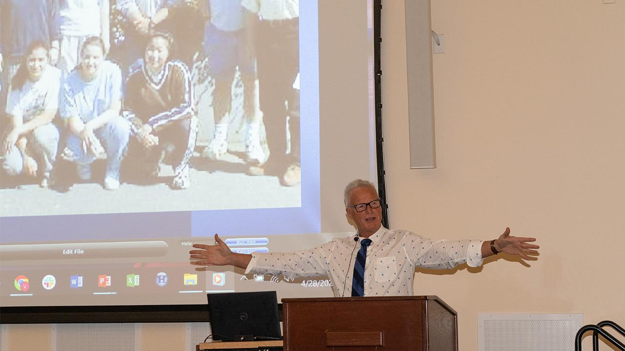 Man talking, his arms spread wide. Above him, a projector screen shows an old photo with a group of students.