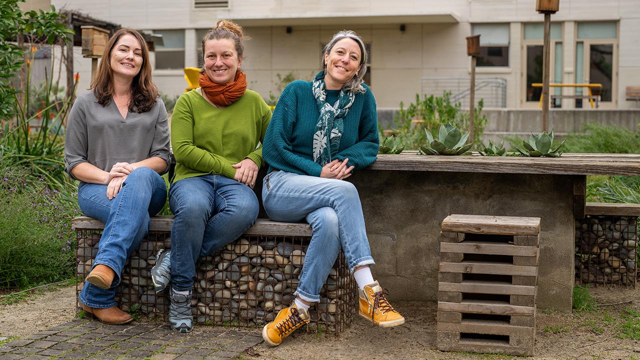 Three women sitting on a bench in a garden. They look happy