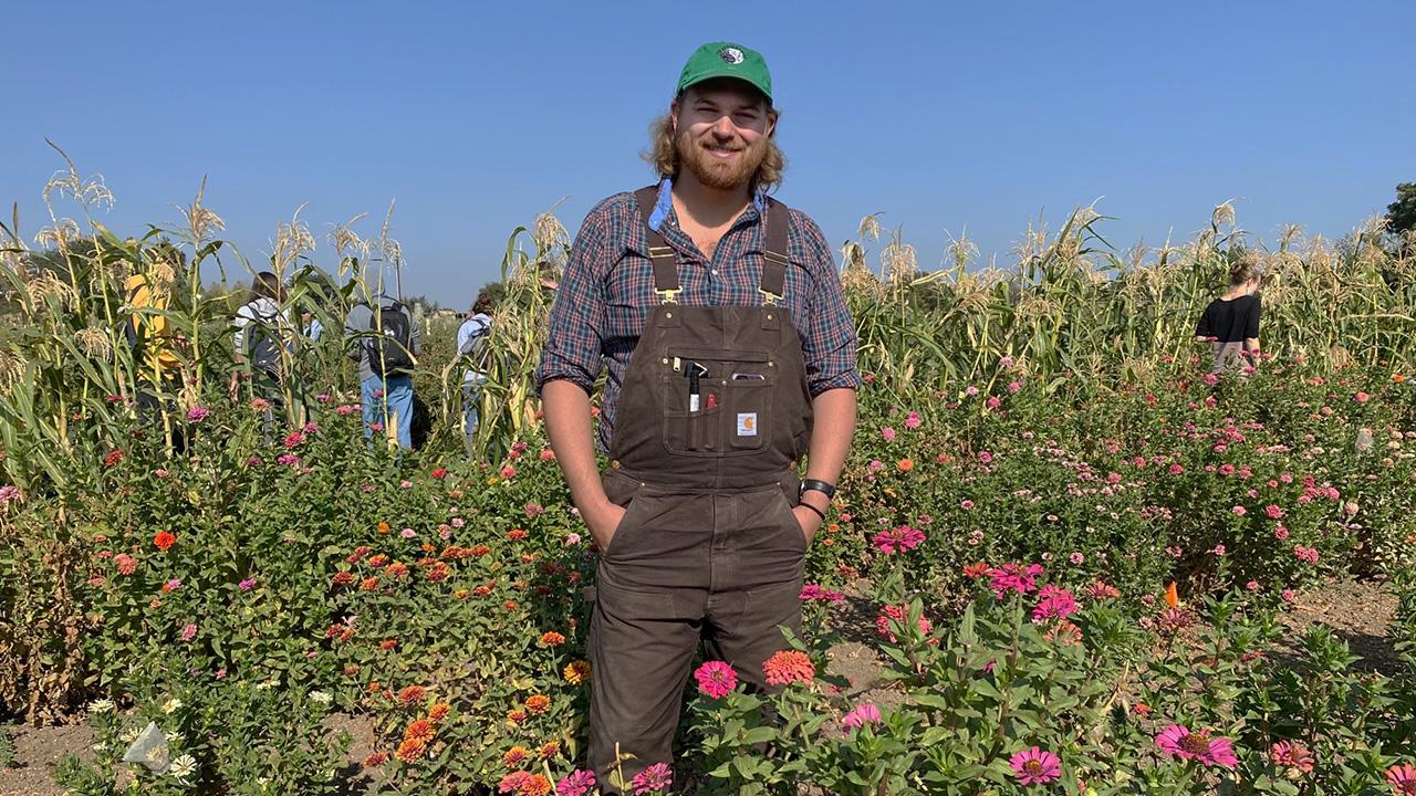 A young man wearing overalls and a ball cap stands among tall bushes bearing pink, orange and wine-colored flowers