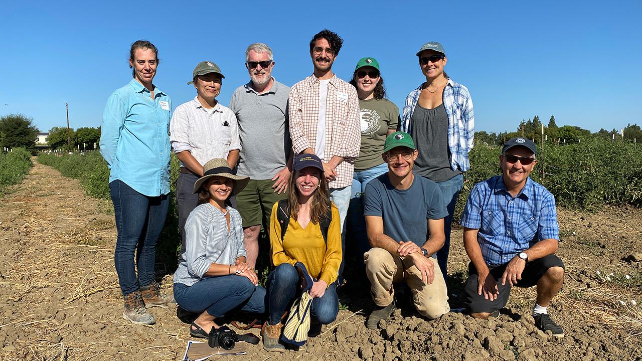 10 men and women in farmwork clothes pose in a field, with a blue sky behind them.