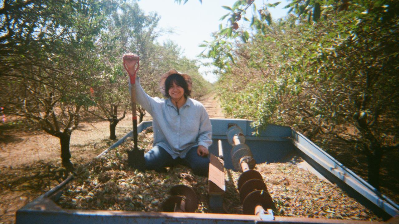 Woman stands on top of wagon in a field of almonds. She is holding a shovel and smiling at the camera.