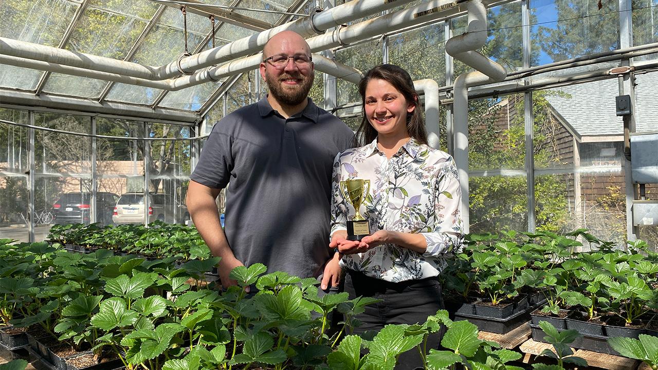 A woman holds a small gold-colored trophy. She stands with a man in a greenhouse. In front of them are trays with hundreds of 4-inch pots with small green plants growing