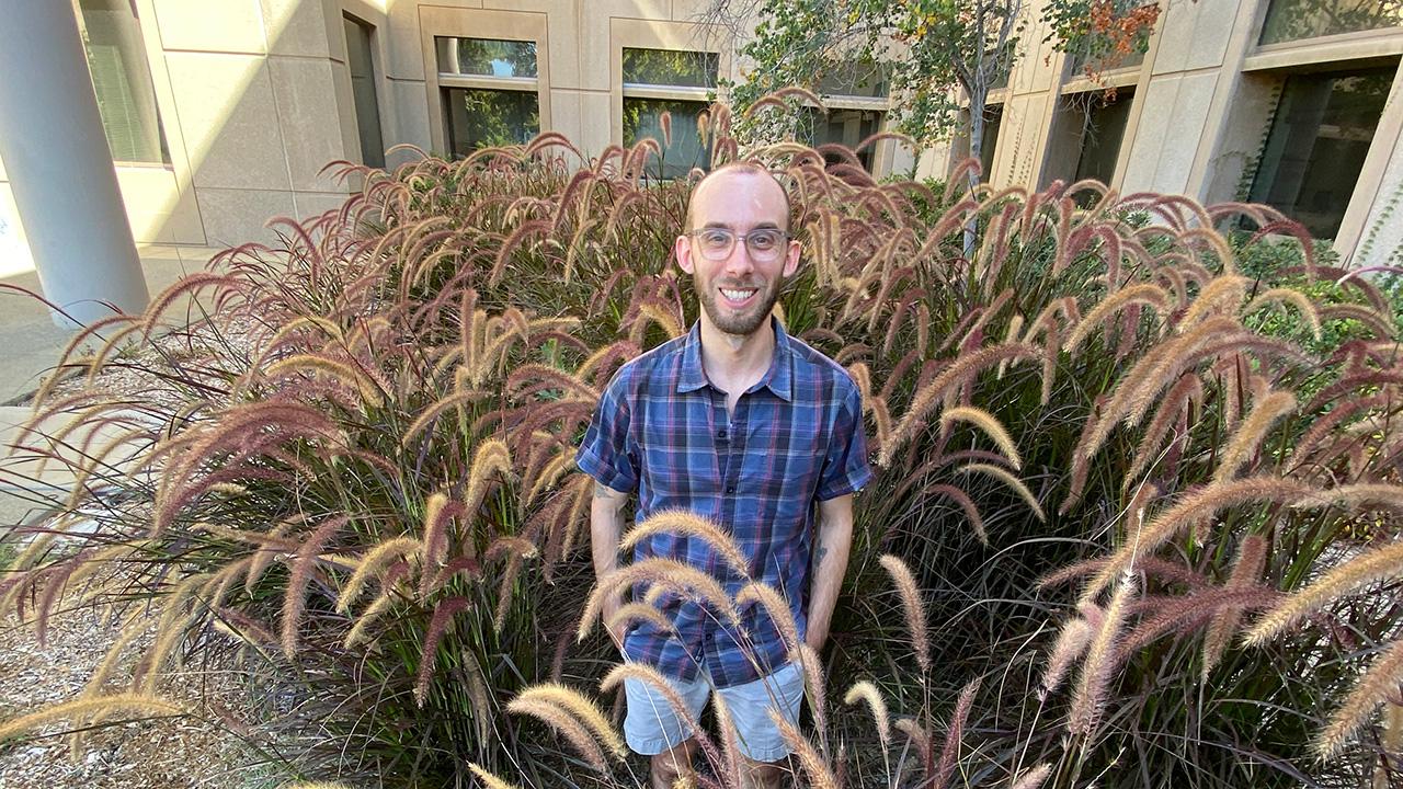 Man standing amid tall bunchy landscape grasses, with a building in the background