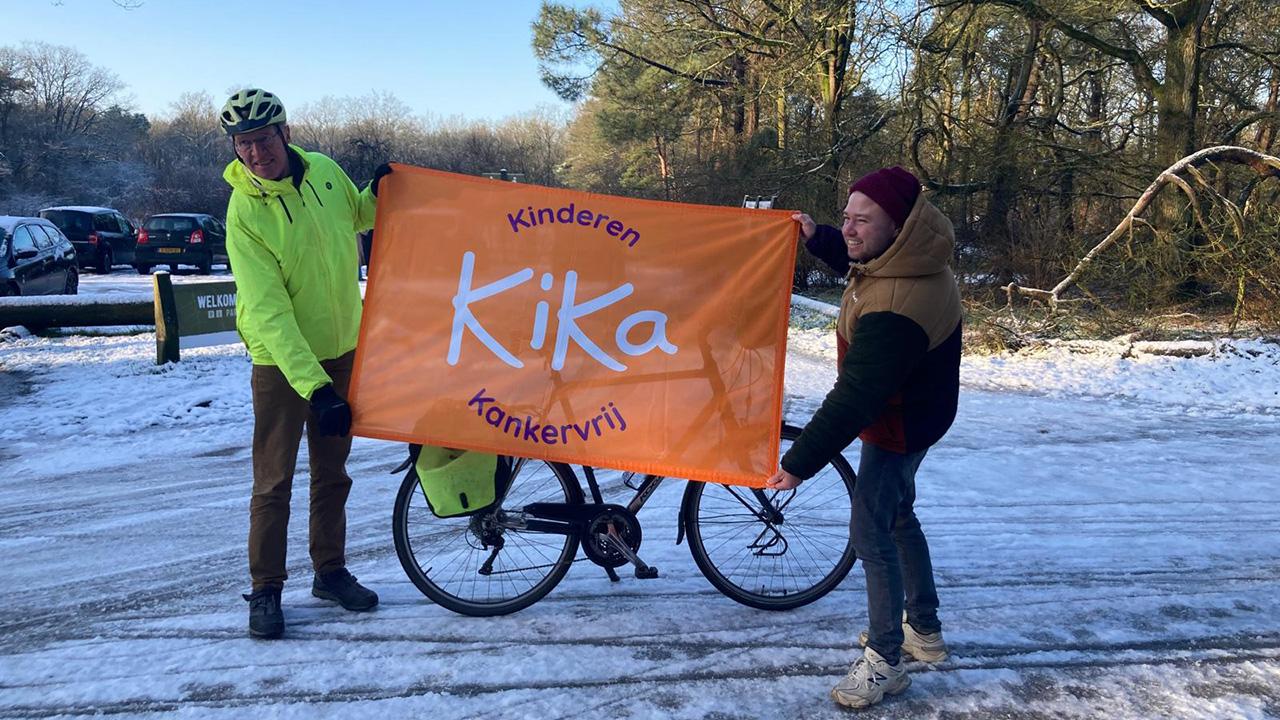 Two men outdoors holding an orange banner. A bicycle stands behind.