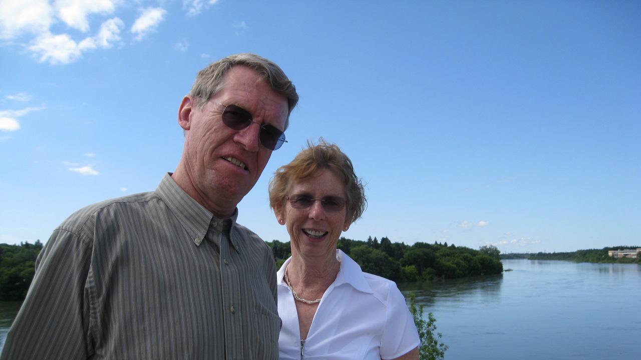 Older man and woman standing by a wide river. Sunny day, blue sky behind them. They are smiling.