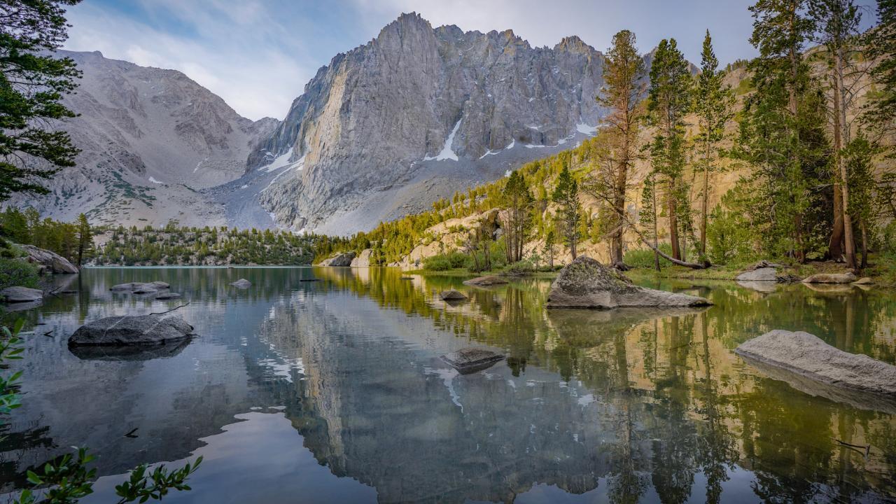 A small alpine lake surrounded by tall pine trees. The exposed rock of a mountain is visible in the background.