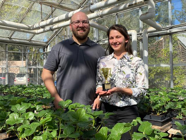 A woman holds a small gold-colored trophy. She stands with a man in a greenhouse. In front of them are trays with hundreds of 4-inch pots with small green plants growing