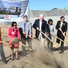People in business attire standing in a field holding shovels and flinging dirt into the air. Behind them are greenhouses behind a fence.