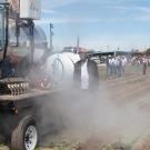 A large, tractor-like machine kicks up a cloud of whitish vapor in a field, while a man stands next to it smiling.