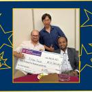 Three young men hold a giant check and are smiling, one gives the thumbs-up