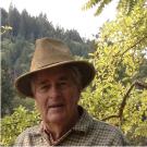 Older man wearing a hat, with a mountainside and trees in the background.