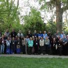 A large group of people standing in 3 rows outside under large trees