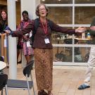 A woman looks joyful, standing with her arms outstretched and smiling in an outdoor patio, speaking to people present.