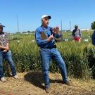 Three men standing in front of tall golden grass. The one in the center is holding a microphone and talking.