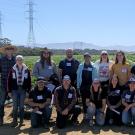 A large group of people standing in front of low furrows, with low mountains and blue sky in the background.