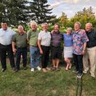 Eight men and one woman stand in a row, smiling, against a backdrop of trees, lawn and cloud-studded sky