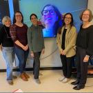 Five women standing in a room. A sixth woman appears on a large TV screen behind them.