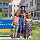 A woman wearing a blue graduation robe and mortarboard stands with two young children in front of a building on a sunny day