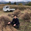 A young man at the edge of a field. Bundles of grass are heaped along the edge, A small truck behind him, and mountains on the near horizon.