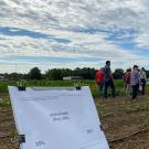 In the foreground, a white sign reads, "glufosinate." It's in front of rows of crops with various levels of looking shriveled or dry. People are wandering among the rows. Above are dramatic gray-and-silver clouds, with a little blue sky showing behind.
