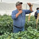 Man with a microphone speaking in a greenhouse-like structure, surrounded by green plants. A few people also visible in the background.