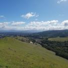 A rolling landscape view from atop a hill looking down into the valley. Trees dot the area with clouds near the horizon.