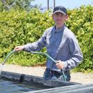 Man standing in front of a hip-high, open, cement water tank, holding a hose and water is running out of the hose into the tank. Grape vines behind him.