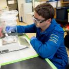 Young man in safety glasses using a lab balance at a cluttered chemistry bench