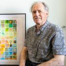 An older man standing next to a large and colorful framed piece of art.