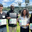 Four smiling young people holding certificates on a riverside lawn with bridge behind them. UC Davis Department of Plant Sciences