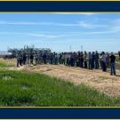 A large group of people by a low field on a sunny day. They're standing on a high area amid the low-lying fields.