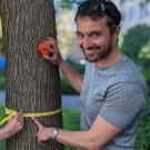 A man and a woman close to a tree trunk, with a tape measure around it. Smiling at the camera.