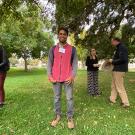 Man standing on grass under large spreading tree, with a few other people nearby chatting.