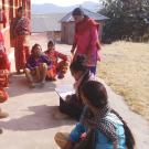 Women wearing colorful red garments, sitting by rustic buildings with the view of high mountains in the background.