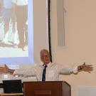 Man talking, his arms spread wide. Above him, a projector screen shows an old photo with a group of students.